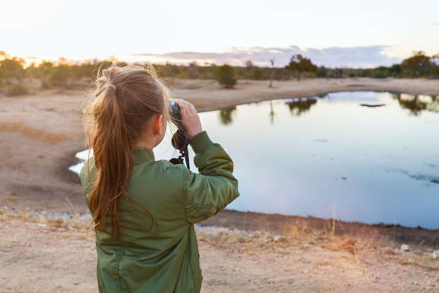 A person with a ponytail uses binoculars to observe a waterhole at sunset, surrounded by dry terrain and sparse trees under a partly cloudy sky.